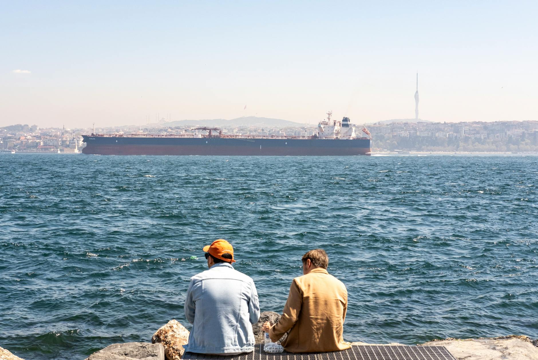Two people sitting by the Bosphorus with a large cargo ship passing, Istanbul skyline visible.
