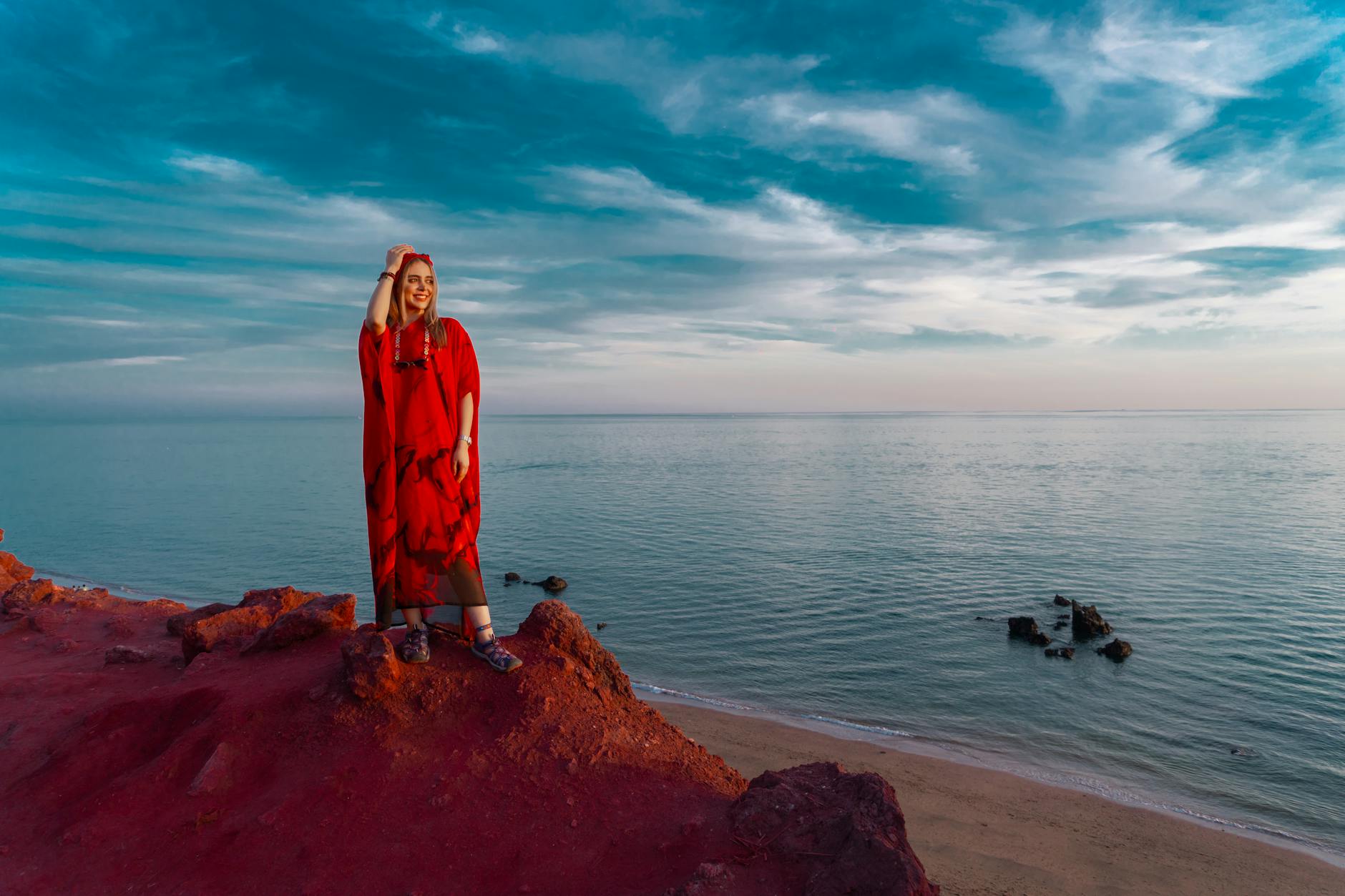 Elegant woman in red dress posing on Hormuz Island's red beach with scenic ocean view.