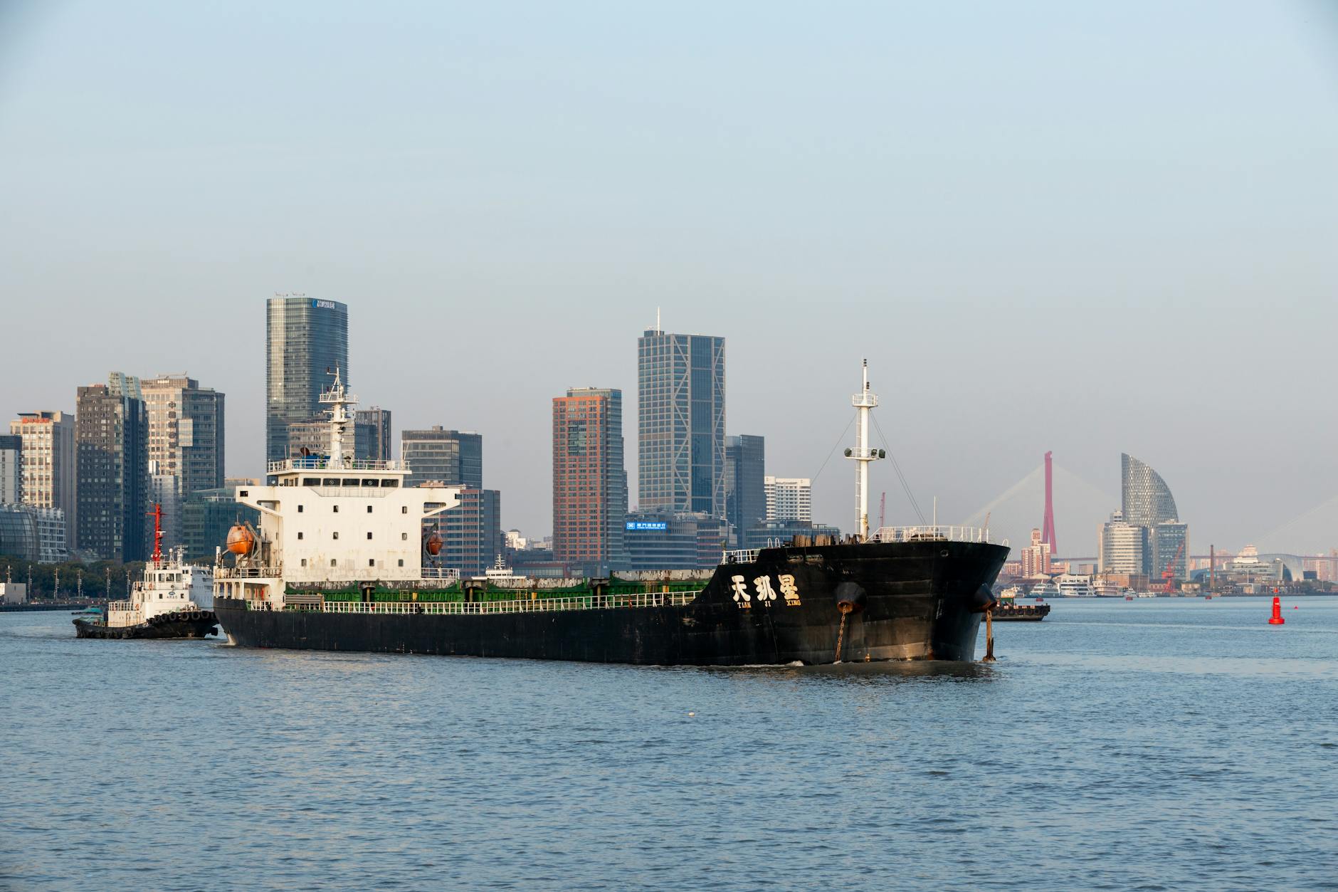 A cargo ship navigates the Huangpu River with Shanghai's skyline in the background during daylight.