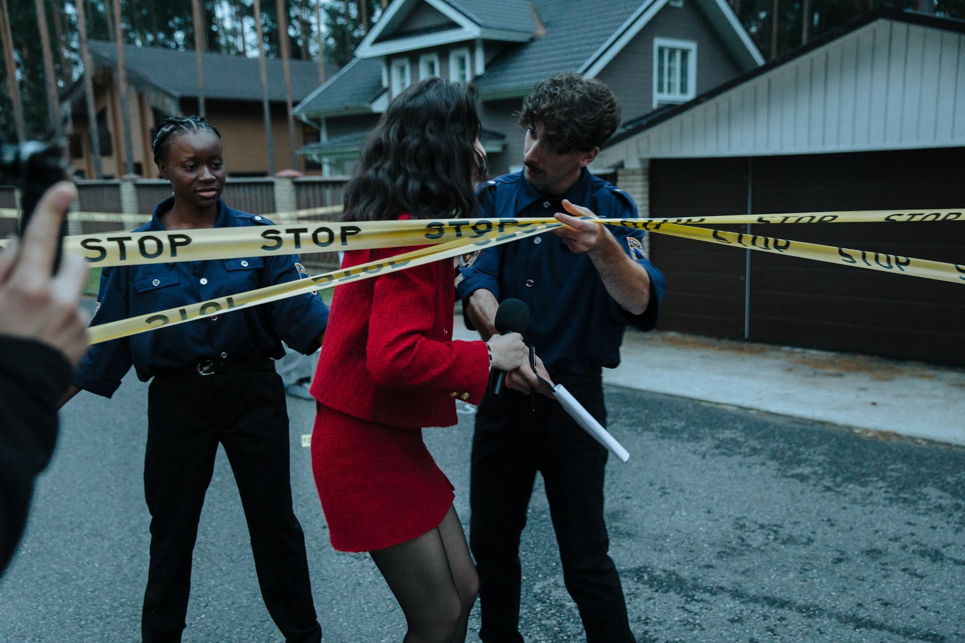 Female journalist interacts with police officers at a taped-off crime scene outdoors.