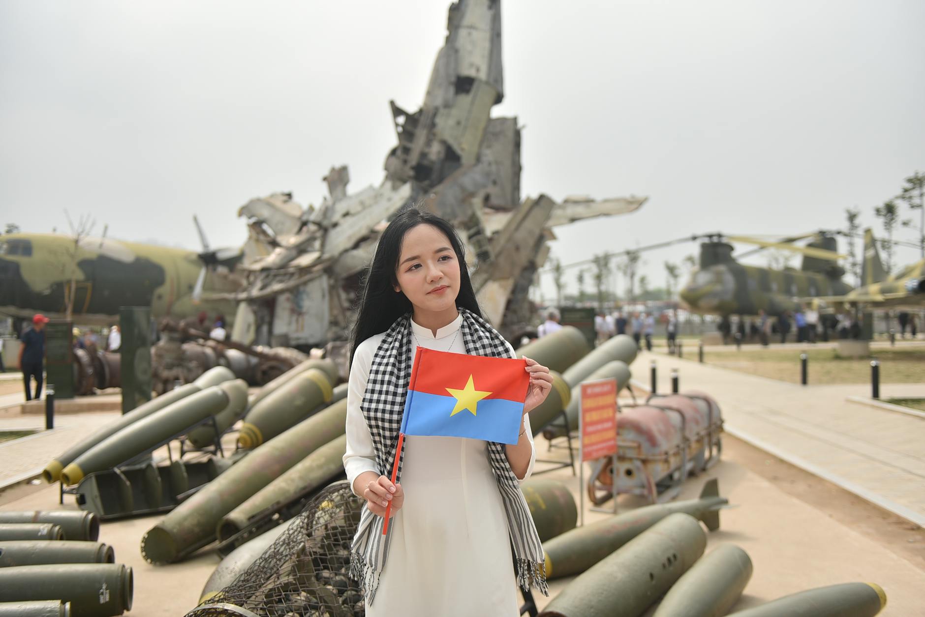 A woman stands with a Vietnamese flag at a historical war museum exhibit outdoors.