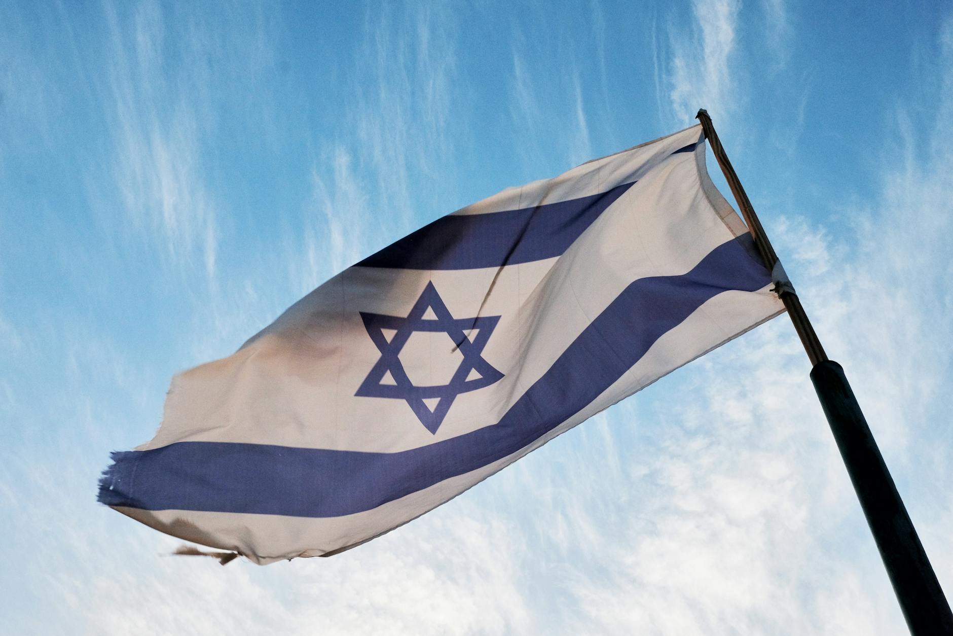 A close-up of the Israeli flag waving on a flagpole against a clear blue sky.