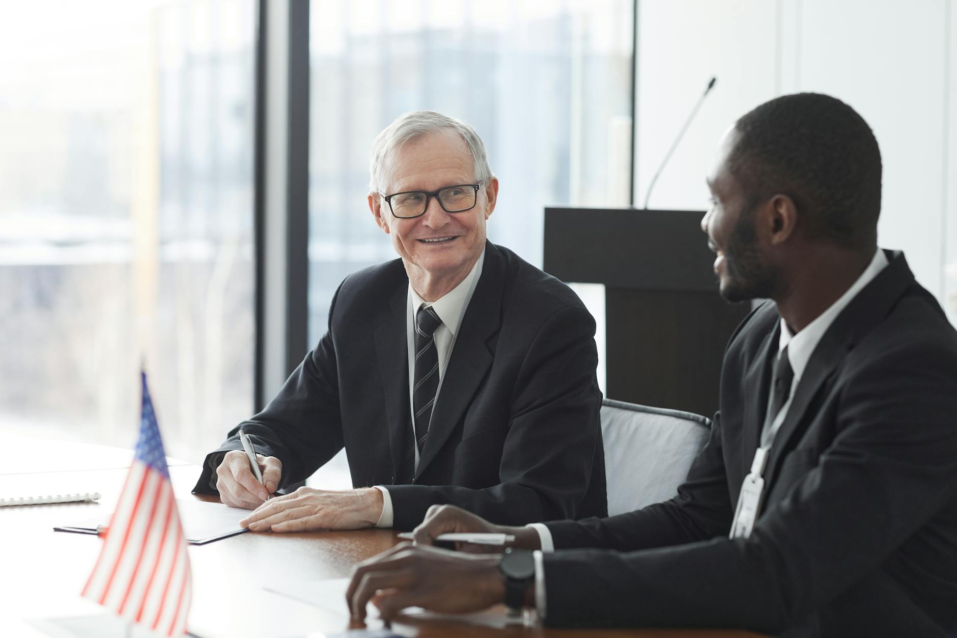 Two business professionals discussing and signing documents at a meeting with an American flag on the table.
