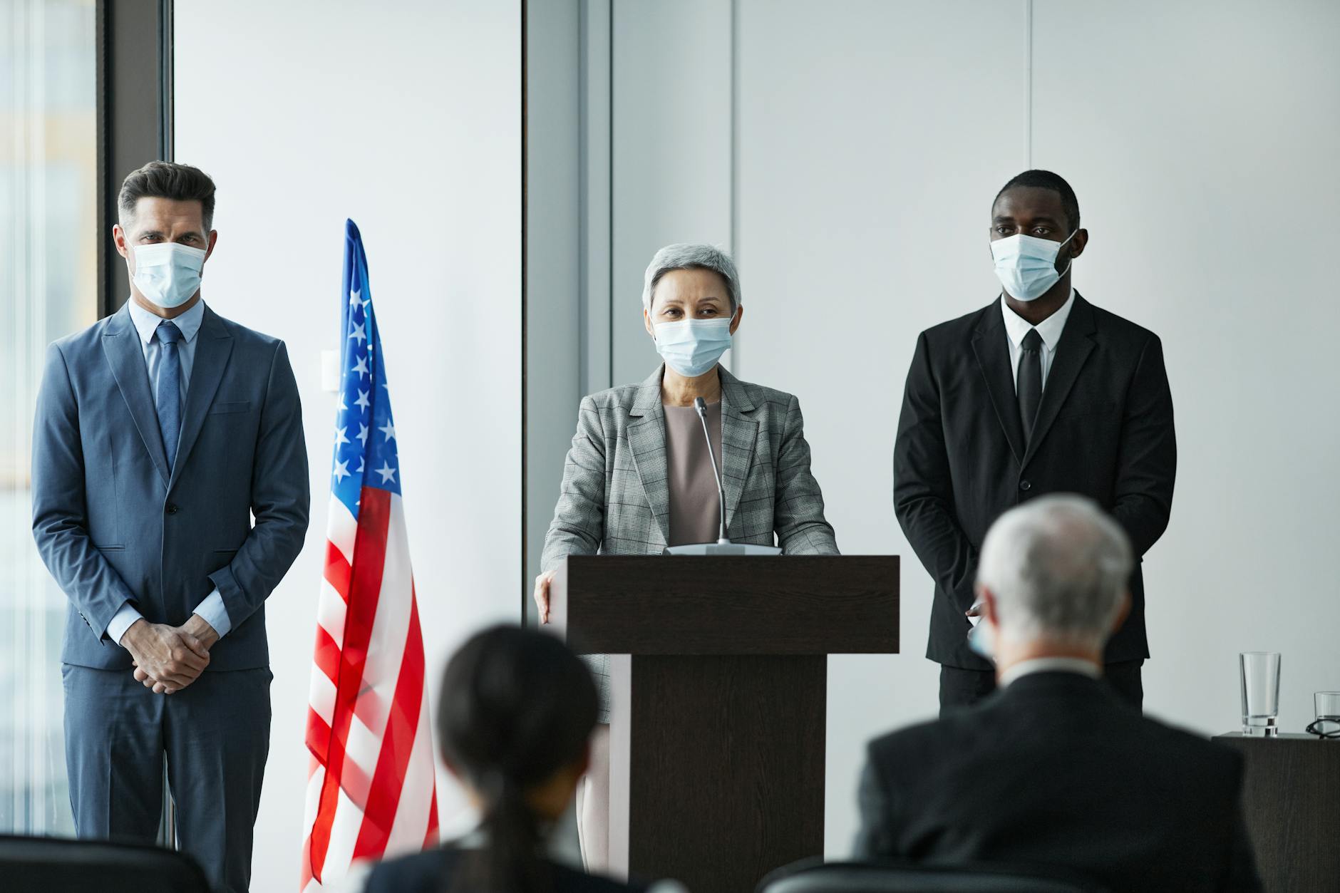 Masked business leaders at a conference podium with US flag backdrop, emphasizing safety and professionalism.