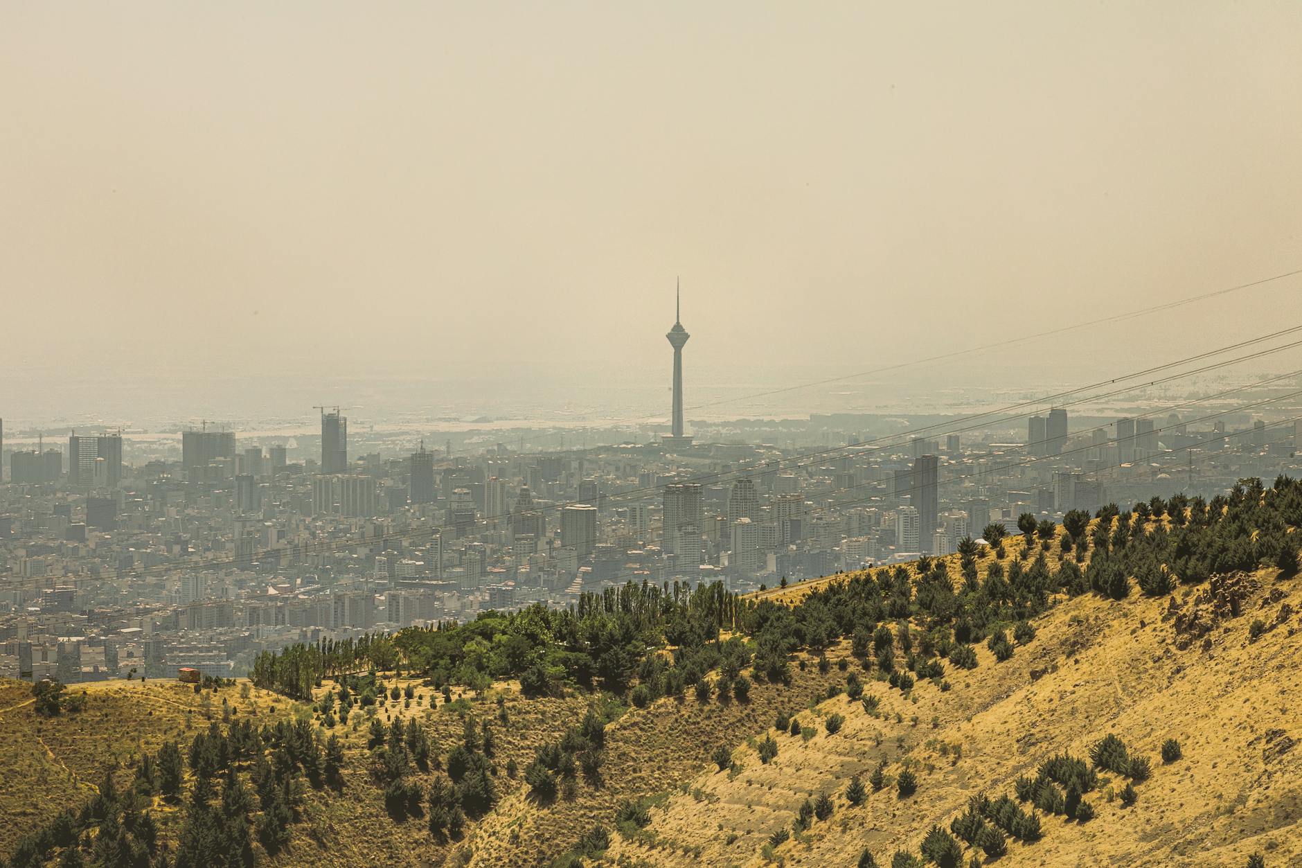 View of Tehran cityscape featuring the Milad Tower surrounded by hills under hazy skies.