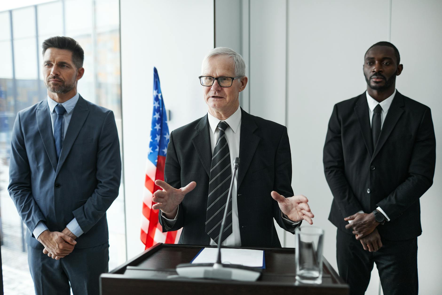 Vance Mockery satire image: Three business professionals giving a speech with an American flag backdrop.