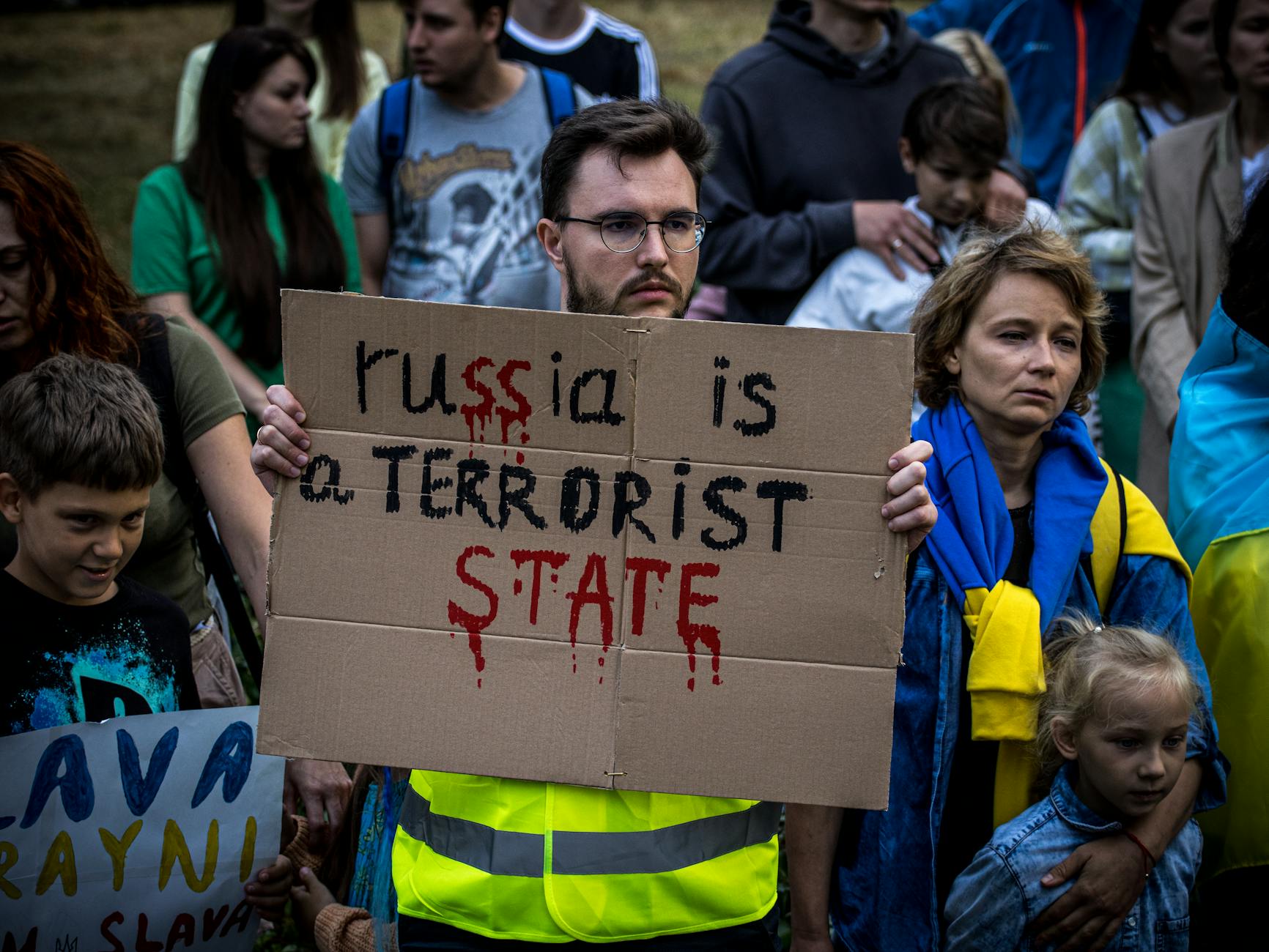 A group of adults and children holding a protest sign in an outdoor rally against Russia's actions.