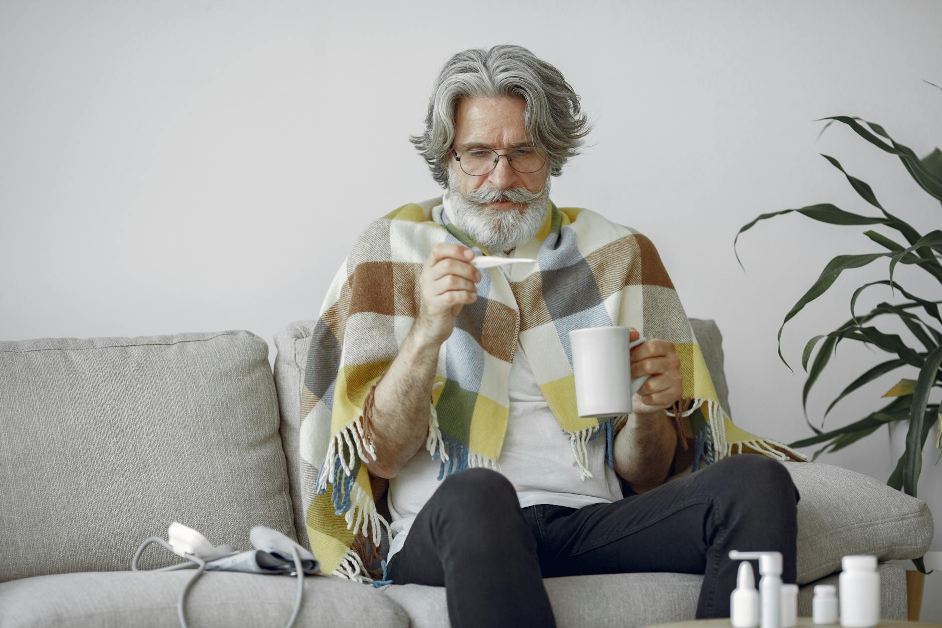 Elderly man checking his temperature while wrapped in a blanket, sitting on a couch holding a mug.