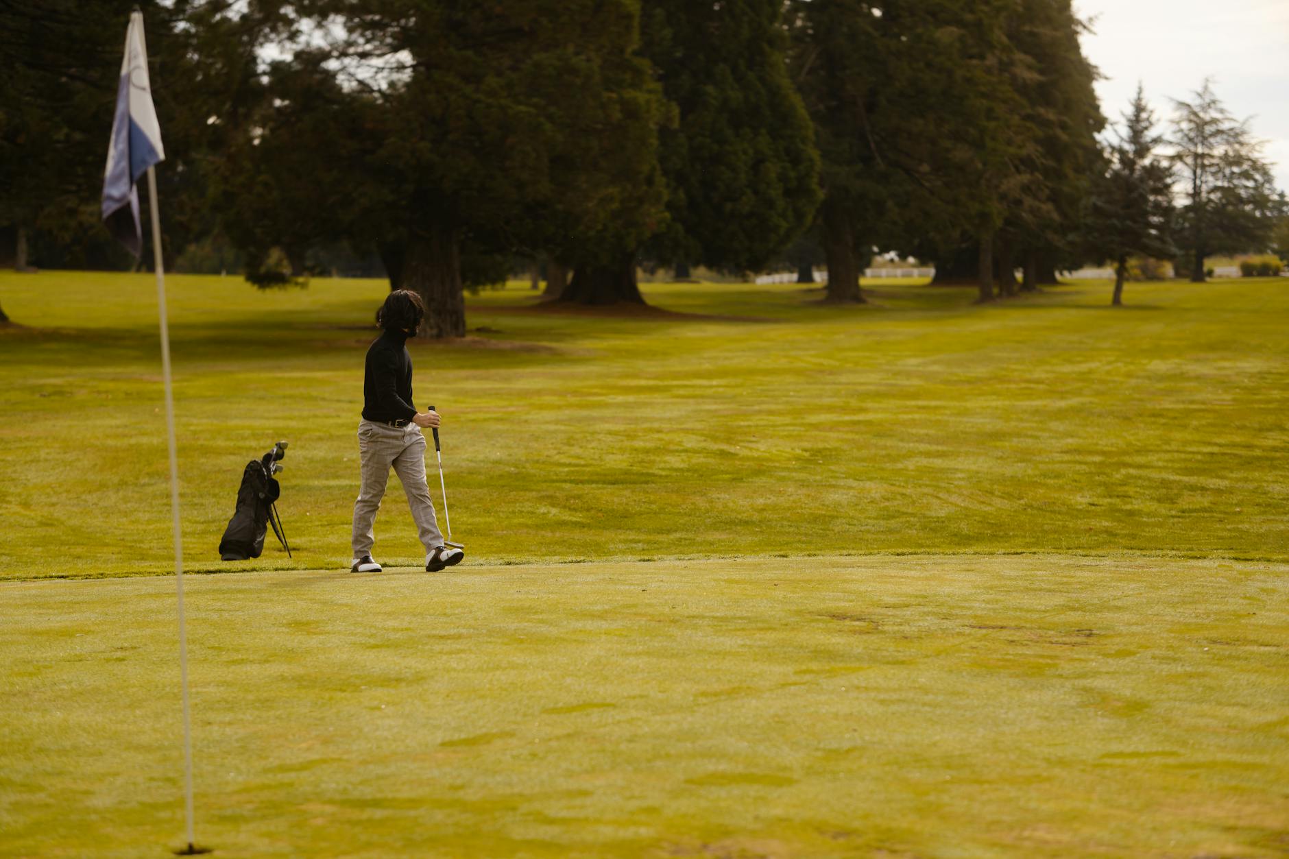 A golfer walks on the green with a golf bag, surrounded by lush trees.