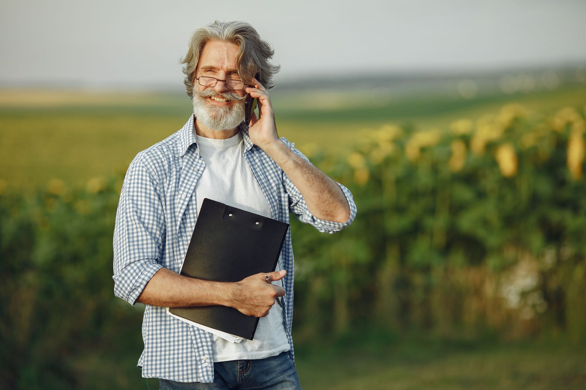 Trump Lowers satire image: Elderly man with beard and glasses talking on phone outdoors in sunflower field.
