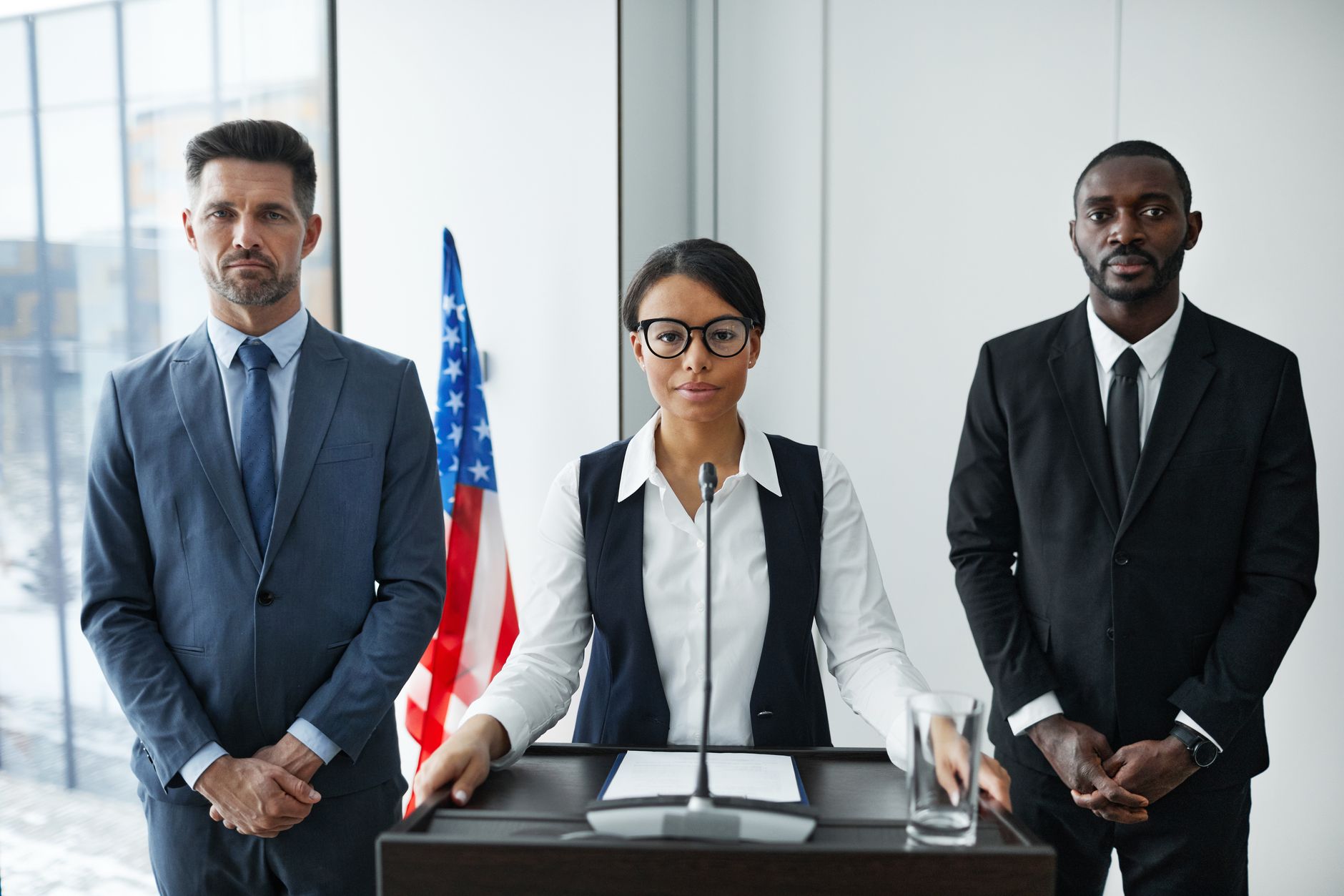 Trump Votes satire image: Diverse group of politicians in suits at a podium with American flag indoors.