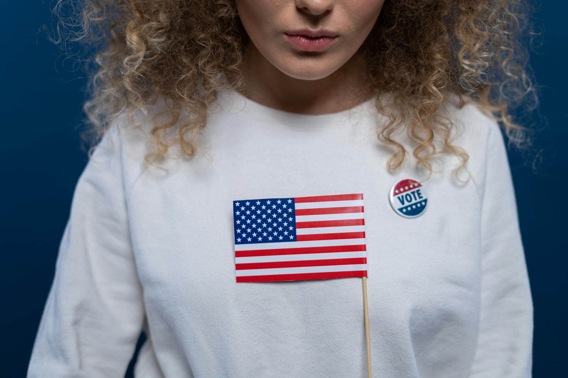 satire-image-23 Trump Republicans satire image: A woman with curly hair holding an American flag and wearing a vote pin against a blue background.
