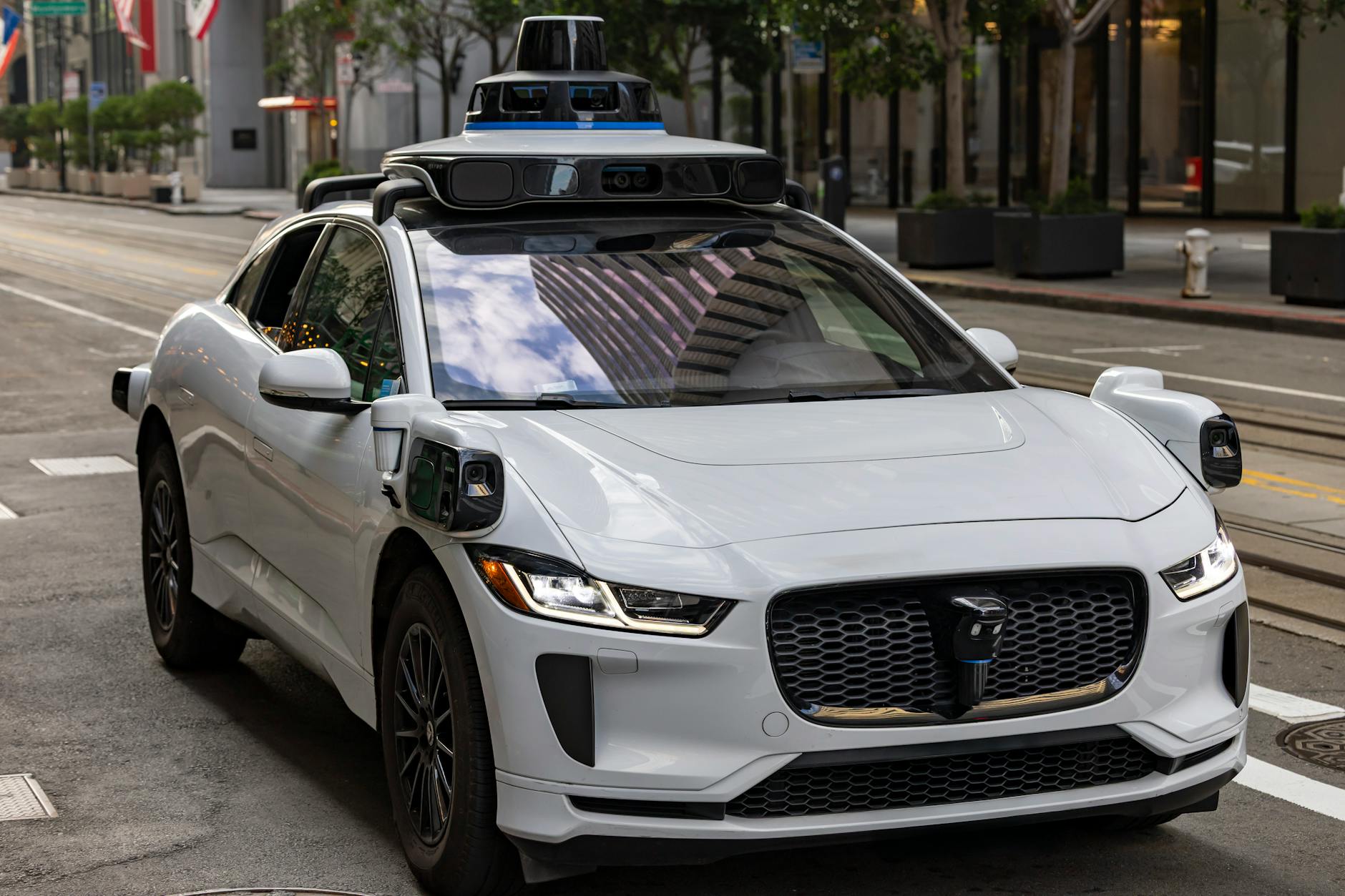 China Demands satire image: A white autonomous vehicle navigating a city street, reflecting urban architecture in daylight.