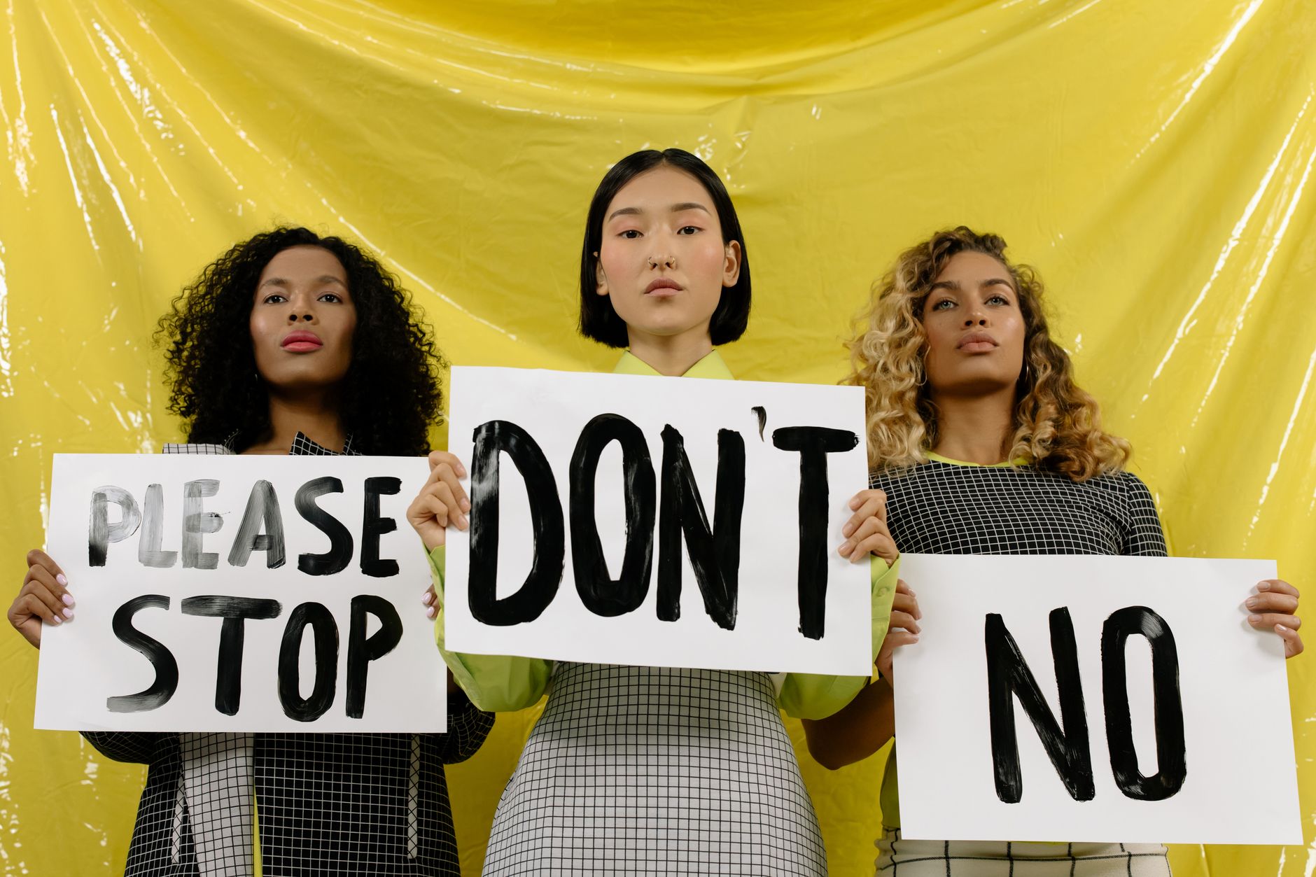 satire-image-61 Trump Marks satire image: Three women stand united holding protest signs against a vivid yellow backdrop.