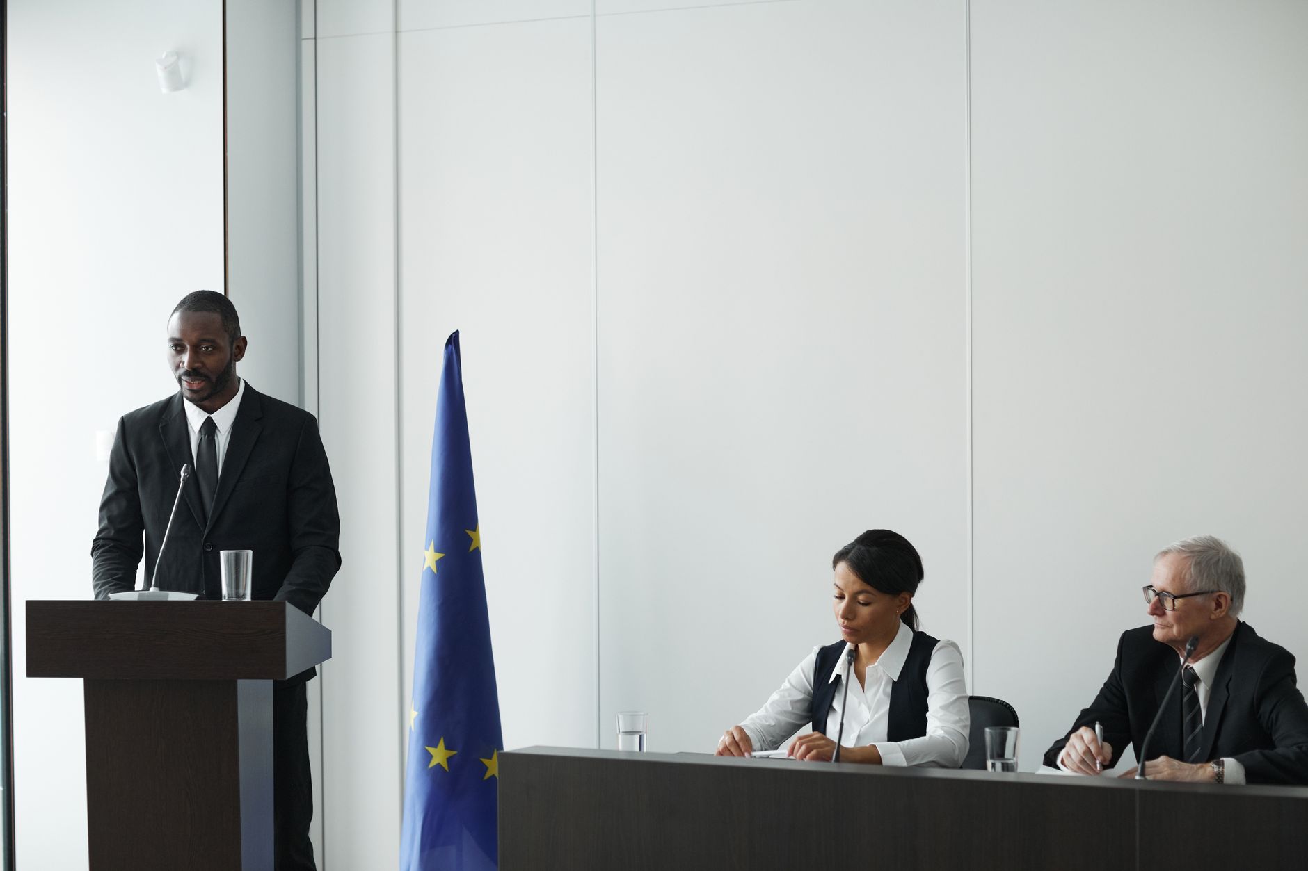 satire-image-63 Trump Installs satire image: Professional business conference with EU flag, diverse speakers in suits discussing diplomacy.